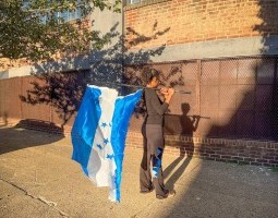 Performer Daisie Cardona, dressed in a black long-sleeved top and black yoga pants with a blue and white patch on one leg, stands facing a building. Seen from behind, she holds the Honduran flag behind her. The flag pole rests on her shoulder.
