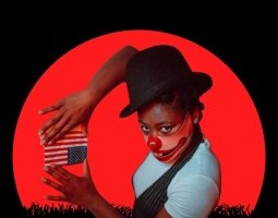 There is a black grassy horizon line and a mostly full red moon rising. In the circle of the moon is a young black woman in a short sleeve white shirt and a black vest. Her Right arm is raised so her hands can frame above and below a small upside down american flag. She wears a circus style top hat and a mask over the bottom half of her face that is a clown's wide smile.