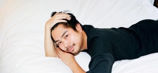 An Asian, male-presenting figure wearing black, lounges on plush white sheets, smiling as he frames his face with his hands.