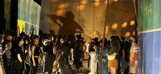 Inside a dimly lit warehouse type space, a crowd of onlookers surrounds 2-3 visible performing artists who wear all black. Green, blue and yellow paintings double as walls. A large shadow of one of the dancers looms above the crowd, projected onto the back wall in warm tones.