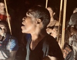 ora chipaumire, a black woman with short hair and the creator of Dambudzo, is in the foreground wearing a black t-shirt and singing into a microphone with her eyes closed as another cast member sings into the background, amidst several audience members.