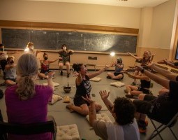 Severin Blake sits on the floor in the middle of a studio space with their arms outstretched toward an audience member. Blake is surrounded by a group of people sitting on chairs and pillows with their arms outstretched as well. There is a singing bowl, some cards, and a broom head sitting on the floor at the center of the space. The studio is dimly lit with two clip lights that are mounted on the wall.