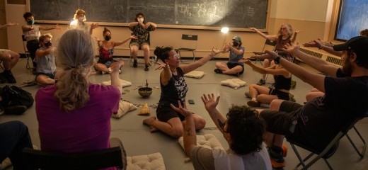 Severin Blake sits on the floor in the middle of a studio space with their arms outstretched toward an audience member. Blake is surrounded by a group of people sitting on chairs and pillows with their arms outstretched as well. There is a singing bowl, some cards, and a broom head sitting on the floor at the center of the space. The studio is dimly lit with two clip lights that are mounted on the wall.