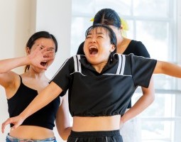 A mischievous joy permeates the image, as one performer with blunt black bangs seems to cheer with an open-mouthed grin, shoulders reaching up to her ears. To her left, another stands gazing at her, with a grin just as wide, but a tad more sinister, an open jazz hand covering her face. Behind the first is a taller person with a yellow bow, tucking something into the first performer’s black-and-white luxury jumpsuit.