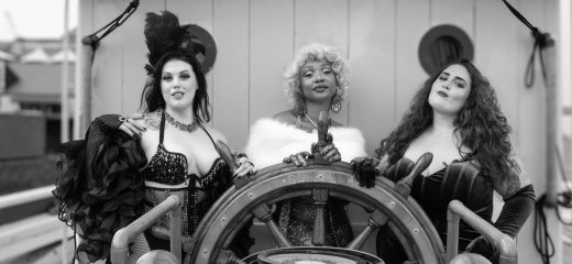 A black-and-white photograph shows three burlesque performers posing confidently at the wheel of Philly's historic ship, the Gazela Primeiro. Regal and composed, AshantiDoll stands directly behind the wheel in a sparkling gown with a white fur stole and short, curled blonde wig. On the left, Raven Nightingale wears a sequined top, fishnet stockings, and dramatic feathered sleeves, accessorized with a tall feathered headpiece. On the right, Miss Angie rocks a velvet corset-style gown with long gloves and voluminous curly hair as she tilts her head with a playful, bold look. The ornate ship’s wheel dominates the foreground,, anchoring the theatrical trio in a maritime setting that contrasts with their vintage burlesque styling.