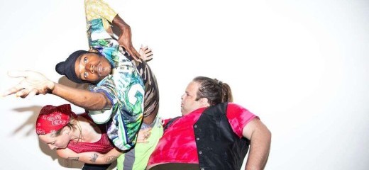 Three dancers in dynamic positions pose against a stark white background. On the left Annie Peterson wears a red bandana and a red pinnie is hunched over looking down. In the center a Vitche-Boul Ra dons a colorfully patterned shirt, has one arm akimbo and the other reaches out towards the lens. On the right Zeze Schorsch in a red tie dye tee with a black vest overlain leans back and looks towards the other dancers.