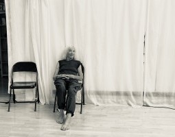 Off center, an old woman sits on a metal chair in front of white curtains. Her hands sit comfortably on top of her stomach, and she smiles mischievously at the camera.