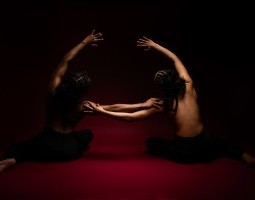 Two dancers sit on a red floor in front of a black backdrop with their hair braided into updos with cornrows. They are topless with their bare backs facing the camera, wearing black leggings. They mirror each other, leaning toward one another with one hand outstretched, resting on the other’s shoulder. The other arm arcs overhead, also in the direction of the other.