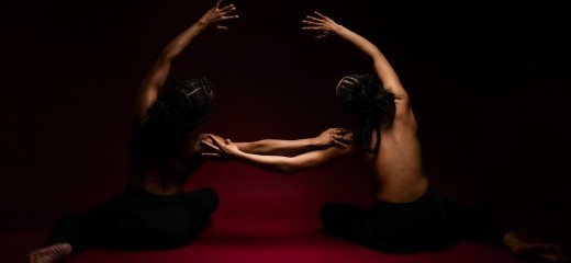 Two dancers sit on a red floor in front of a black backdrop with their hair braided into updos with cornrows. They are topless with their bare backs facing the camera, wearing black leggings. They mirror each other, leaning toward one another with one hand outstretched, resting on the other’s shoulder. The other arm arcs overhead, also in the direction of the other.