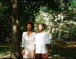 Two Black performers stand side-by-side, wearing pedestrian clothing – lacy, patterned white tops layered over shades of beige and brown. Their gaze extends out to their right, converging on an unknown entity in the distance. Surrounding them, a park with green grass and trees foregrounding urbanity.