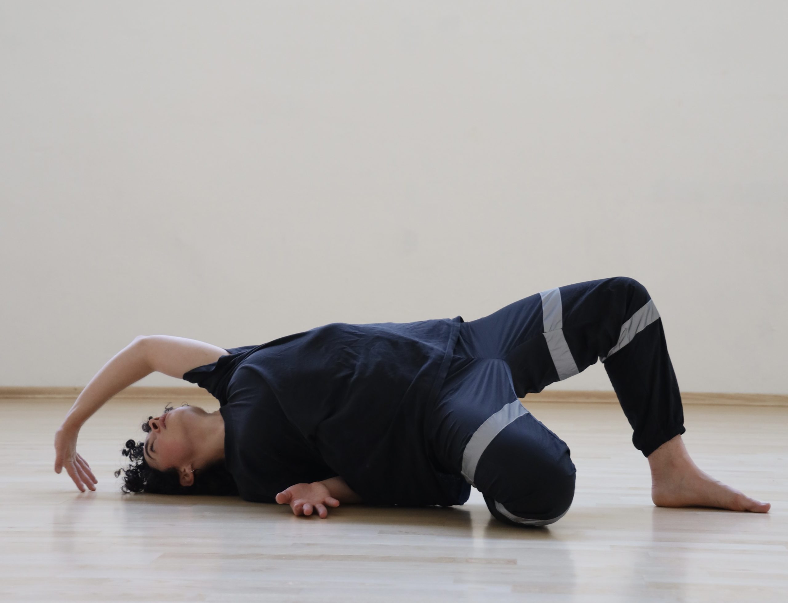 Donovan moves across the floor on hands and feet, emphasizing grounded momentum and fluid contact. He is wearing a soft navy blue shirt and pants and dancing on a  light wood floor in front of a white wall