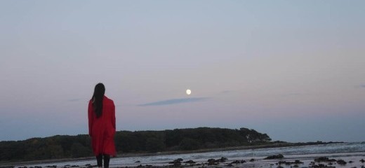 View of Nichole Canuso from behind standing on the beach, her left foot in mid-footfall, at night in an orange coat with the full moon high in the sky.