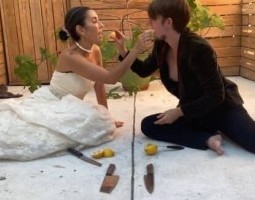 A bride and groom sit on white concrete barefoot with knives and lemons in front of them, feeding each other lemon wedges with a fig tree in a pot and wood fencing in the background.