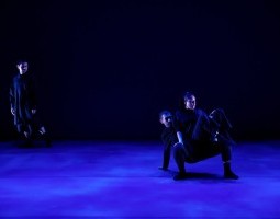 A stage bathed in blue light, performers all in black. One stands at the left side of the image, glancing center-stage at two dancers. Those two dancers morph together into one as one-crouches, belly up in a crab-position balanced on one hand, and the other sits precariously on their lap, knees up to their chest.