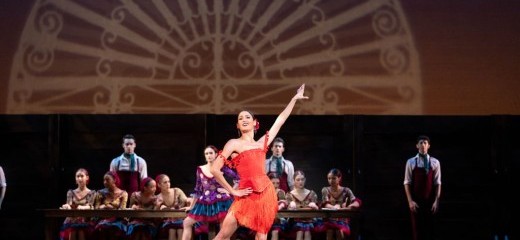 Mayara Pineiro of Philadelphia Ballet performs in Carmen, choreography by Angel Corella. She wears a bright red dress, with one hand affixed to her hip and the other extended outwards on the diagonal plane. A group of dancers sits at the table behind her.