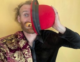 A white person with curly hair, a beard, and piercing blue eyes shows half of zir face, covering the rest with a red dome shaped hat. Pain au chocolat is stuffed in zir mouth, and zir clothes are bifurcated, much like zir face––half outfitted in red and gold, and the other half in black.