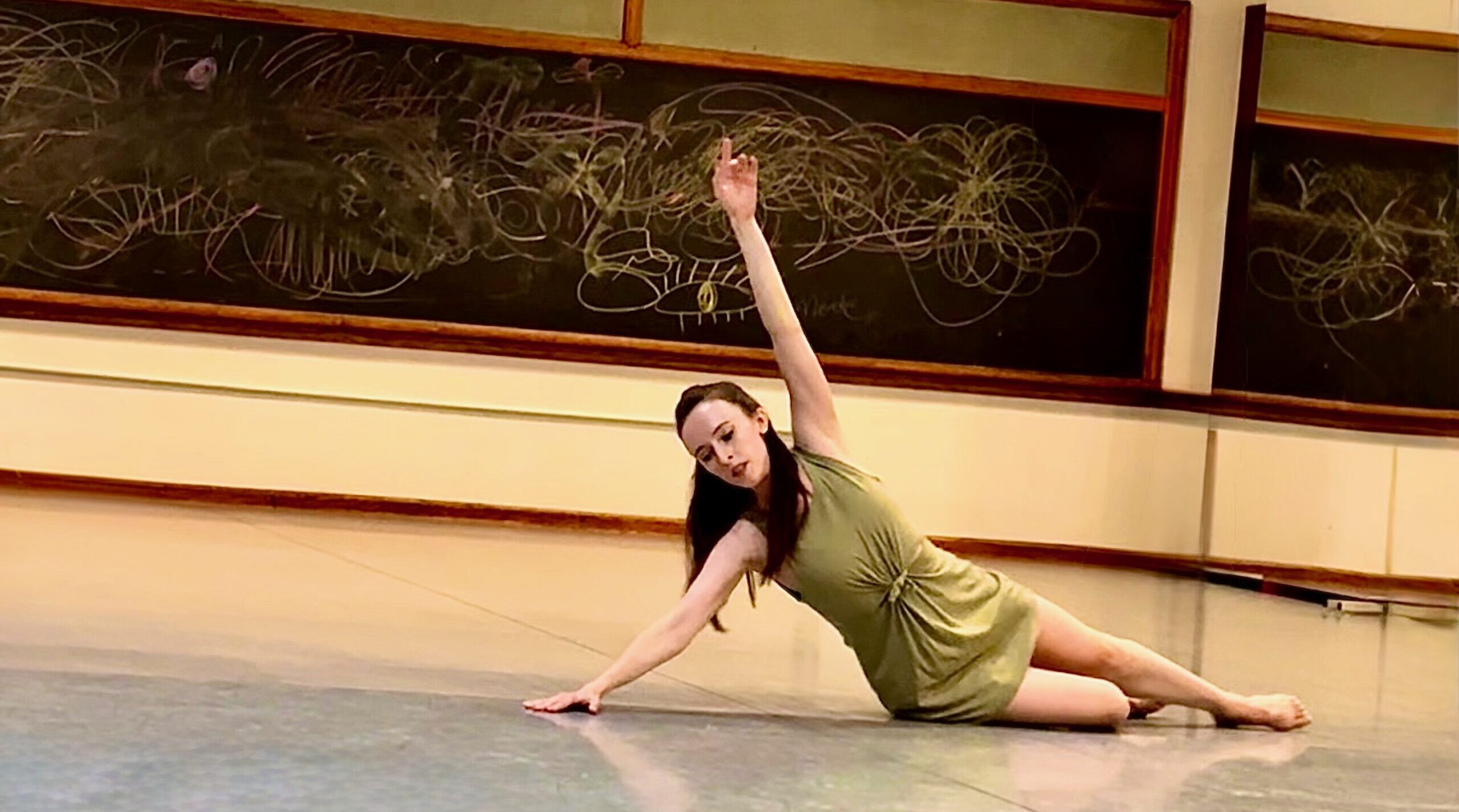 Marina Kec, hair down and barefoot, slides to the floor. A chalkboard covered in hand-drawn loops stands in the background.