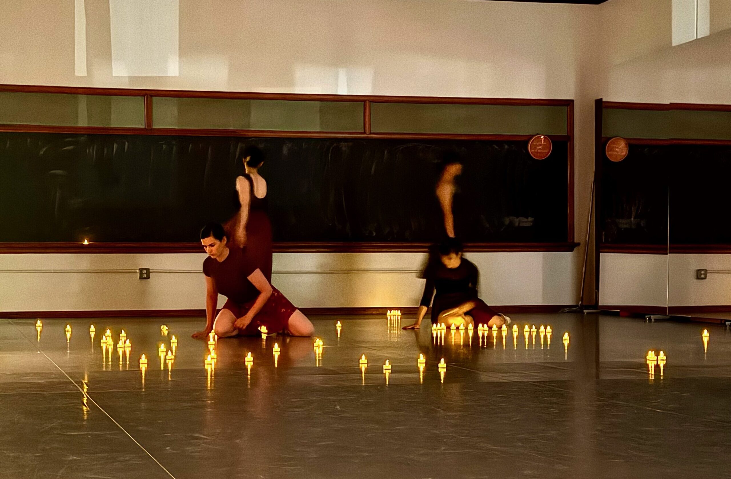 Four dancers move among scattered votive candles in a darkened studio, the studio lights off, their forms illuminated only by the flickering flames.