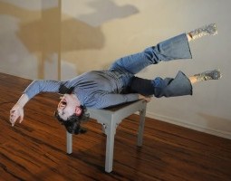 A white woman with dark brown hair, wearing a blue outfit, sparkly shoes, and a bright red lip that matches her red hoop earrings, lies back on a small gray table. Her mouth is wide open as if singing, one arm reaching emphatically above her head while the other grips the table tightly for balance. Her legs float in the air, parallel to one another at a slightly awkward angle. She is a rockstar in an empty room.