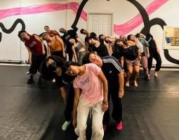 A large group of dancers stand closely together in a studio, their bodies tilted in unison with heads falling dramatically to one side, creating a strong visual wave across the room. They are arranged in staggered rows that draw the eye from front to back, emphasizing their collective rhythm and connection. The black floor grounds the image, while the wall behind them bursts with bold, curving black outlines and sweeping pink brushstrokes, giving the space an energetic, almost theatrical backdrop.