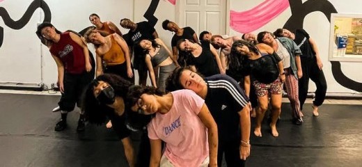 A large group of dancers stand closely together in a studio, their bodies tilted in unison with heads falling dramatically to one side, creating a strong visual wave across the room. They are arranged in staggered rows that draw the eye from front to back, emphasizing their collective rhythm and connection. The black floor grounds the image, while the wall behind them bursts with bold, curving black outlines and sweeping pink brushstrokes, giving the space an energetic, almost theatrical backdrop.
