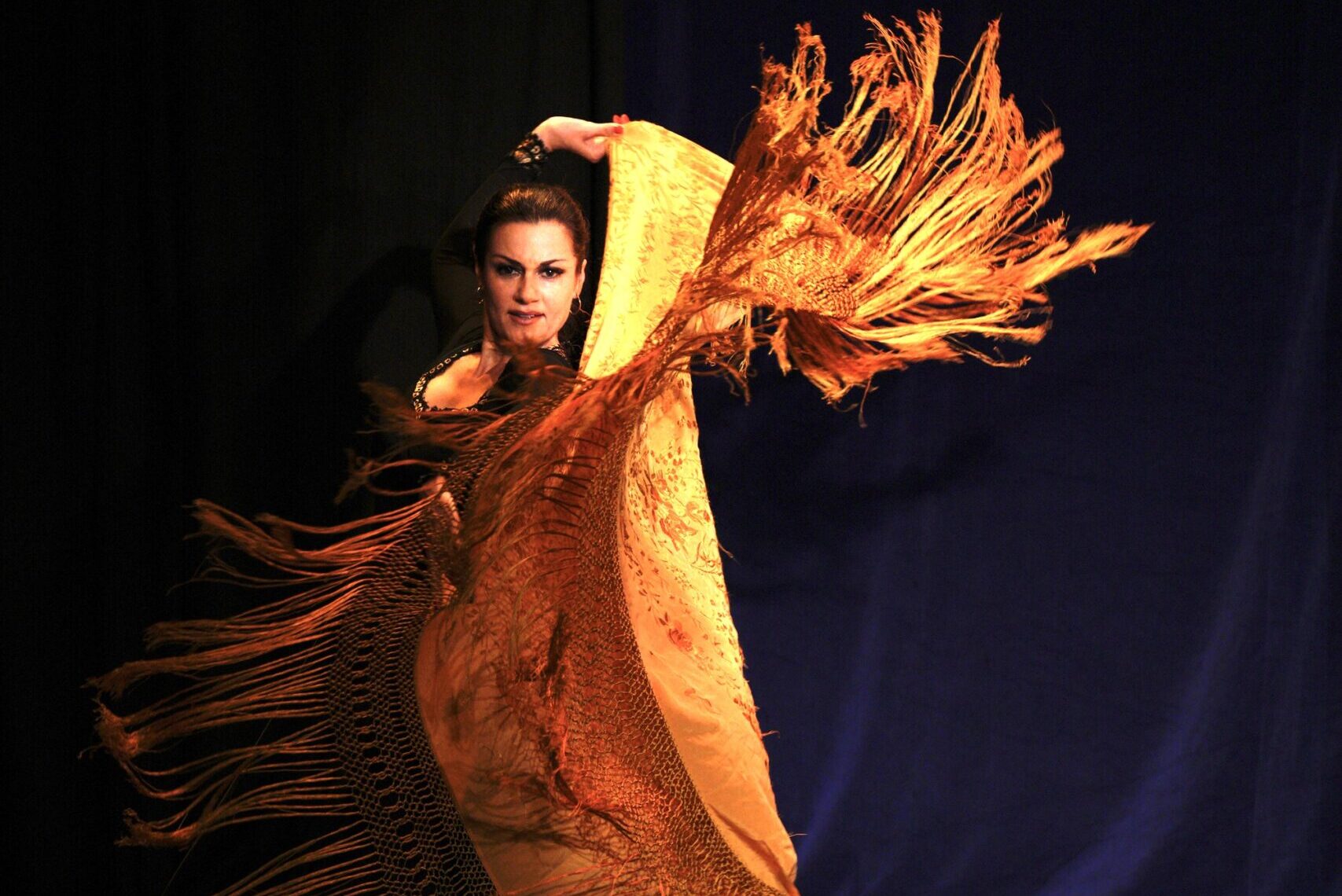 Dancer Aylin Bayaz looks directly into the camera as she lifts one arm above her hand, holding onto the golden tasseled bottom fabric of her dress. Her black hair is slicked back. She looks both triumphant and joyful. 