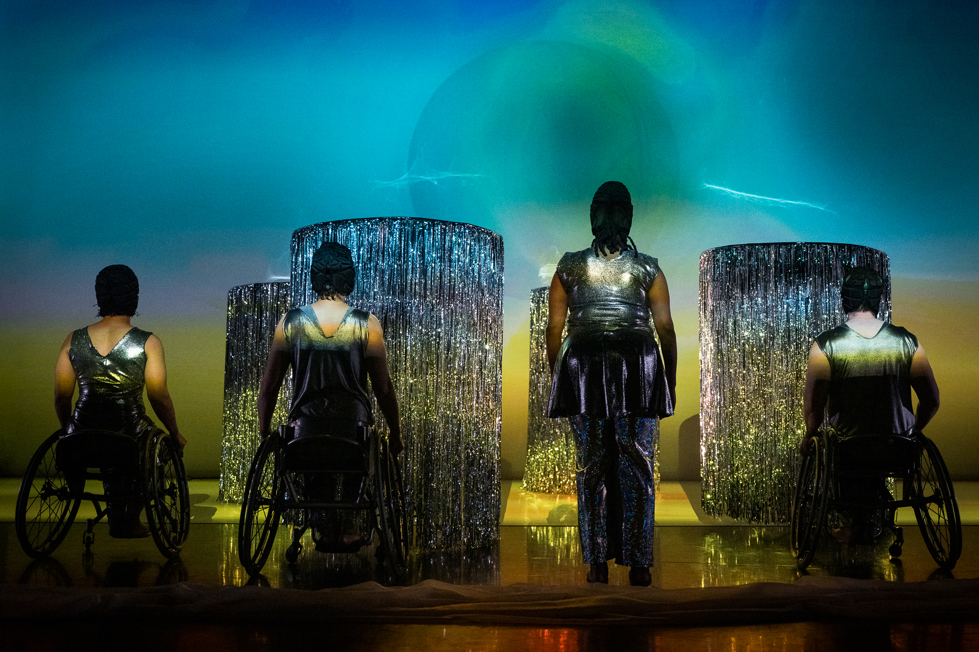 Four performers dressed in silver, with black caps, pause with their backs to the audience, three in chairs and one standing. They gaze at four shimmering towers of silver fringe.