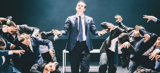 Wearing sharp pantsuits, a group of approximately one dozen dancers from Netherlands Dans Theatre & Complicity lean away from one man in the middle, hair pomaded, gesturing directly to the audience.
