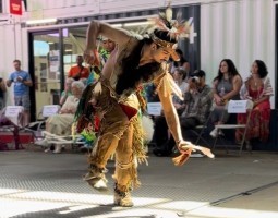 A man dances, hunched over with one arm punching downwards. He wears a headdress and decorative costume elements that highlight traditional artistry.
