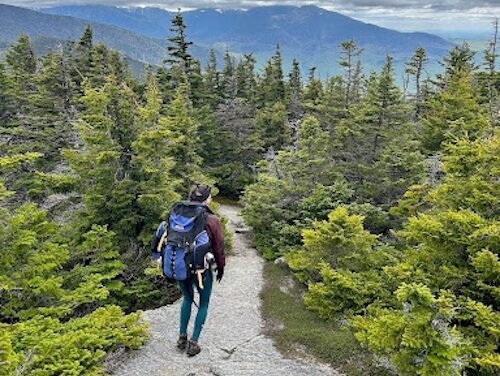 Two thousand feet below rolling gray clouds, a woman walks away down the rock slab trail off the summit of Mount Moriah. She traipses between squat, sturdy, coniferous trees in her fluffy brown zip up, teal leggings, and worn hiking books. A water bottle threatens to tumble out the outer right pocket of her blue Kelty backpack. The backpack has seen some things; it is nearly 20 years old.