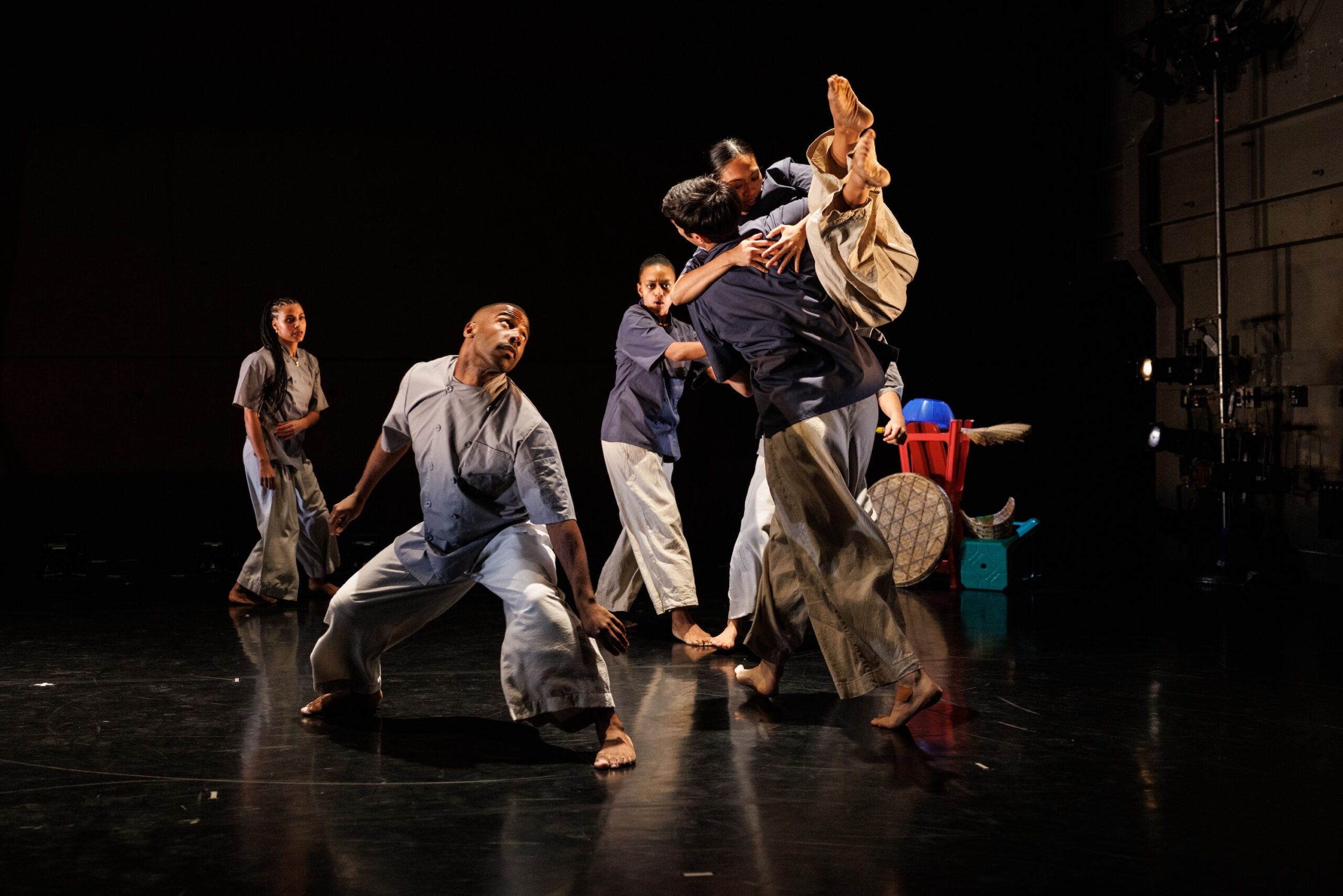 The Of Dishes and Dreams cast in blue Thai-style garments used for restaurant work. Eyes focus on choreographer Keerati Jinakunwiphat as the ensemble walks around her. Keerati is being lifted into the air in a hug, legs piked together, reaching up to the sky. On the left side of the image, Claude “CJ” Johnson, holds another point of focus – legs open in a wide stance, bending deep at the knees, shoulders and hips to one diagonal, while he peeks playfully toward Keerati in the opposite corner. Kitchen items made of plastic and twine balance precariously in the background.