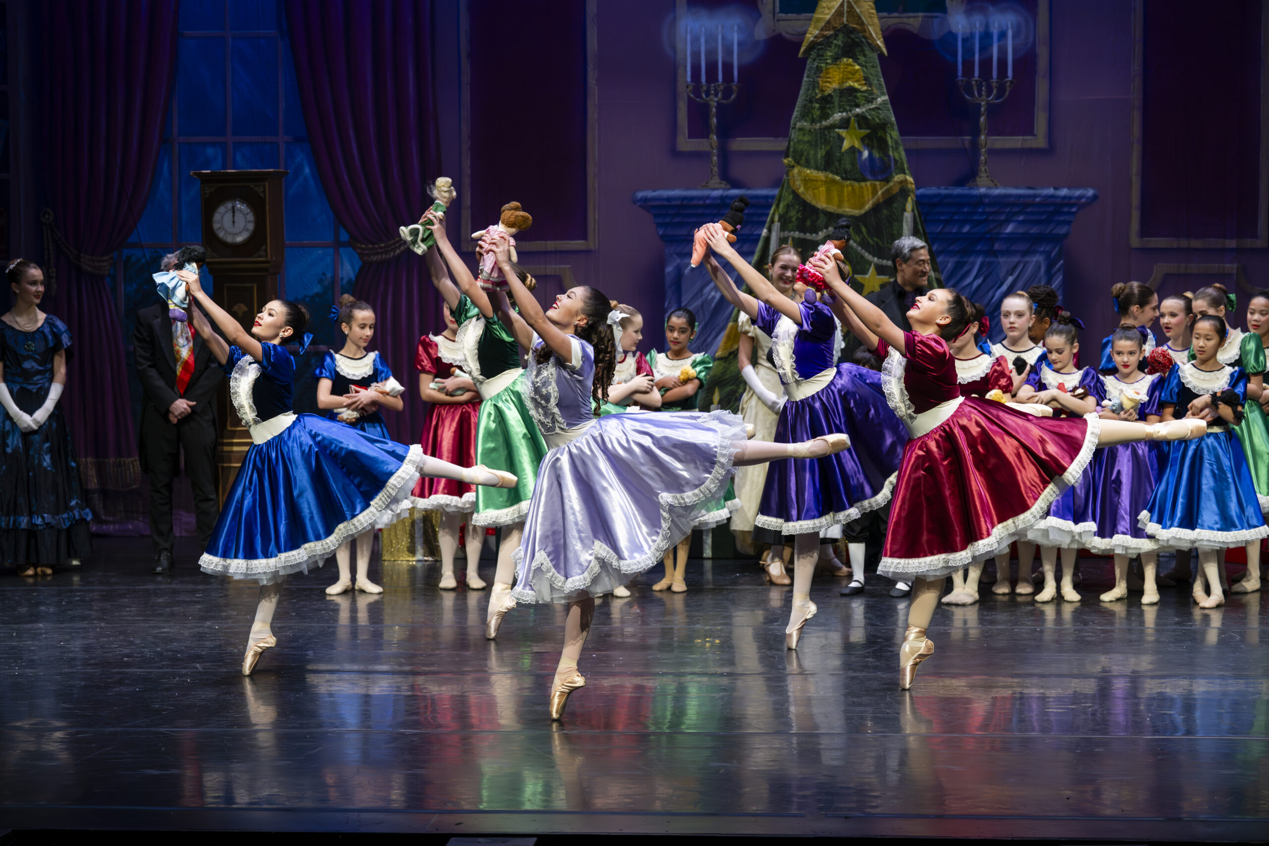 Three dancers from Philadelphia Dance Academy wearing long, bright, billowing dresses, lift dolls into the air en pointe, their left legs extended in arabesque. A group of happy onlookers stand behind them.