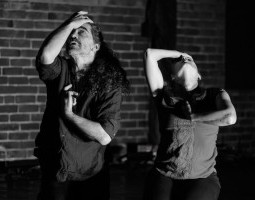 A black-and-white photo of two dancers in a brick-walled room. One, masc-presenting, has long curly hair and peeks out at the ceiling, mouth slightly open in expressive thought, one hand bent to touch their forehead, shielding half of their face. The other hand rests against the center of their body. A second dancer stands to their left, mirroring this pose with face tilted all the way to the sky and taut arms.