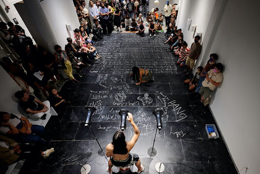 Image Description: The photo is taken from the balcony, looking down on the audience clustered in a U-shape against and besides the white gallery walls, around the black chalk stage, where Baldoz is kneeling to scrawl on the chalk and Vo is standing, legs wide, behind the center of three black megaphones. Some of the audience is shadowed, people stand, or sit on the ground. From this angle, Vo is in the very bottom of the photo, the top of their head, arms, and their bare back visible under a soft spotlight. With white chalk, Baldoz has written in huge letters and small, at all angles, and in a mix of cursive and print. One word, “HANOI”, is legible even from the balcony view.