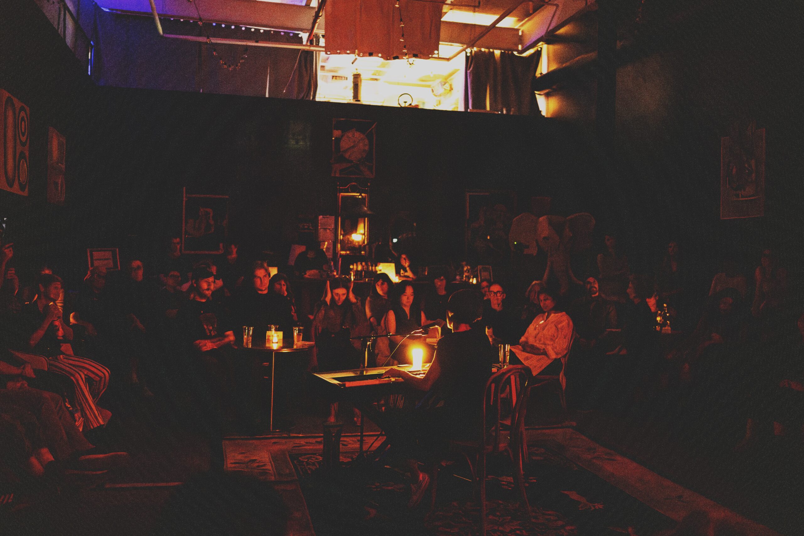 A woman sits with her back to us while sitting at a keyboard. A candle on top of it illuminates the faces of about fifty people who sit facing her in a dim orange color. On the shadowy walls we can see framed paintings, a clock, and an artificial head of an elephant.