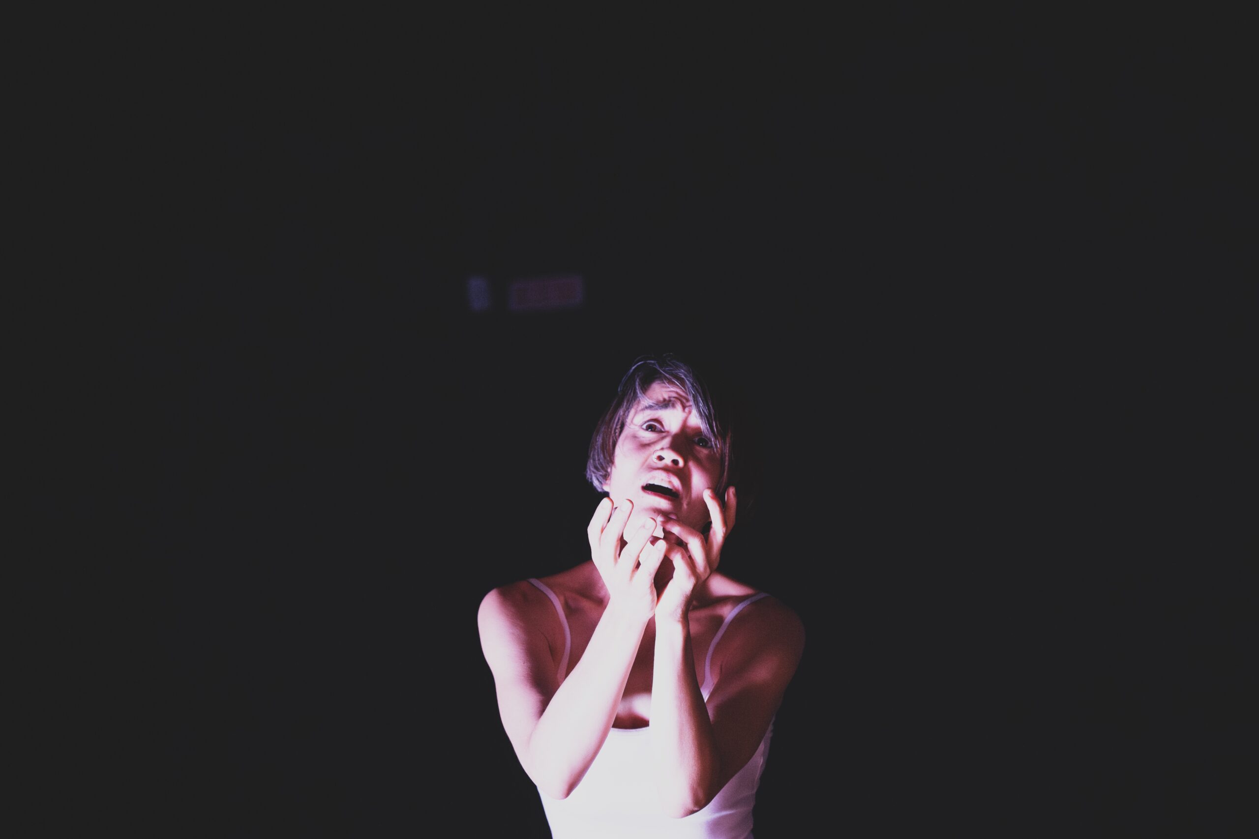 An Asian woman with short greying black hair faces us, the space behind her dark and empty. Her face looks longingly at us, almost in anguish, and their hands extend towards us as if to cup our face, or to beg for mercy.