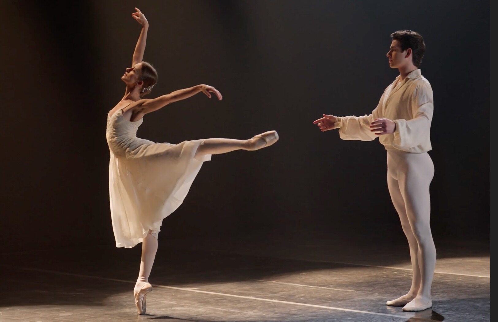 Oksana Maslova balances on pointe in first arabesque, leaning back toward Sterling Baca, who stands behind her in support. She faces upward, arms extended, with a poised, expressive posture; both dancers’ faces convey stoic grief.
