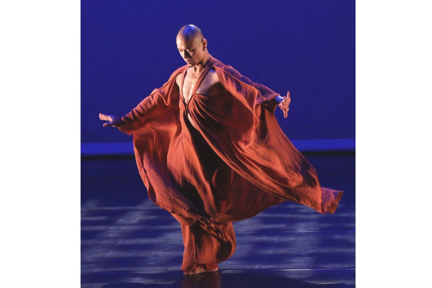 Male dance artist Kun-Yang Lin balances on one leg in flowing red silk, hands flexed gracefully, looking downward while holding a poised dance pose.