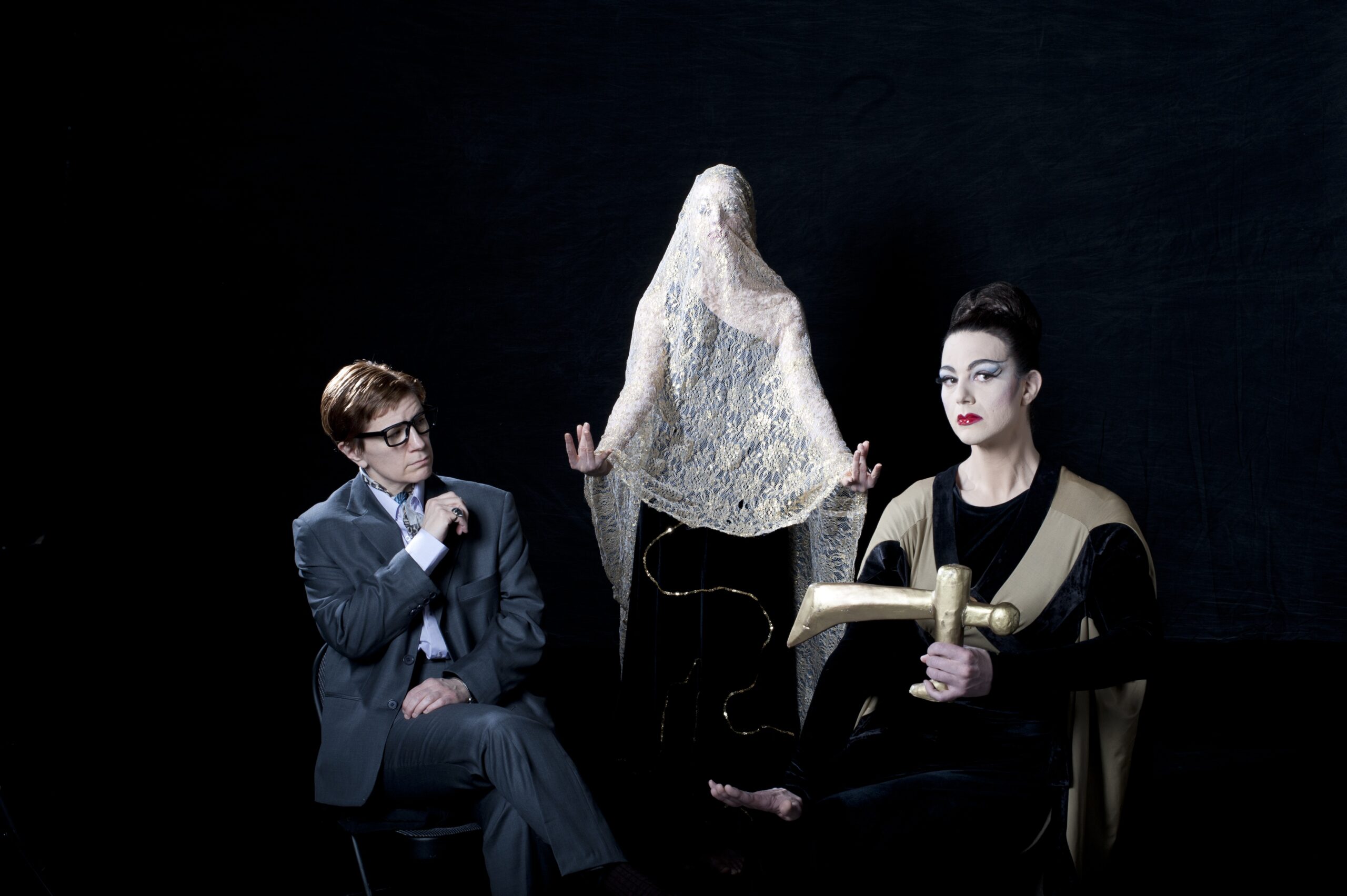 Lisa Kron, playing dance critic Walter Terry, has short brown hair, wears a dark suit and thick-rimmed glasses, and sits with their legs crossed and their arms in front of them. Opposite sits Richard Move, transformed into dance icon Martha Graham: wearing dramatic white make-up and eyeliner, their hair up in a bun, their hand holding a mysterious object that looks like a primitive cross. Between them, a woman wearing a black dress stands with her arms outstretched, a white veil covering their head and torso like a ghost.