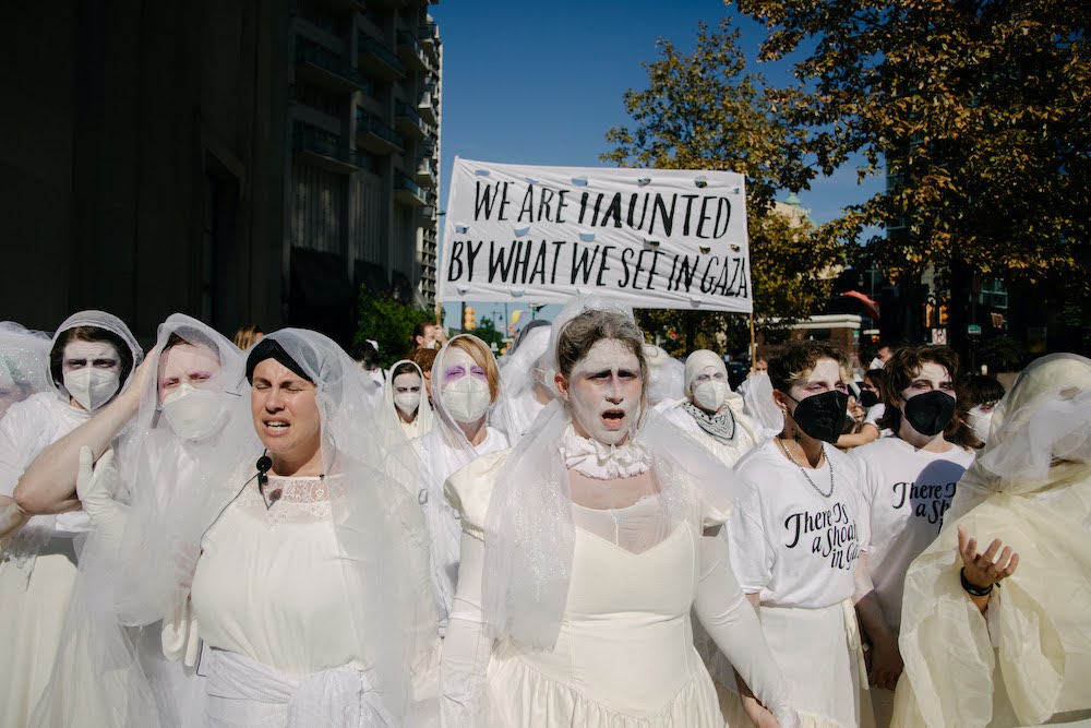 A crowd of predominantly white people wear white dresses, t-shirts, and gauze veils draped over their heads, faces visible. Most have painted their faces white with pink around their eyes and are wearing KN95 masks. They are walking forward toward the camera, a tall shadowed building, tree foliage, stop lights, and a square of blue sky behind them. Two people in the front open their mouths mid-song and scrunch their eyebrows together, maybe from the sun and maybe with concern. Deeper into the crowd, people hold a white banner with bold black letters, “WE ARE HAUNTED BY WHAT WE SEE IN GAZA.”