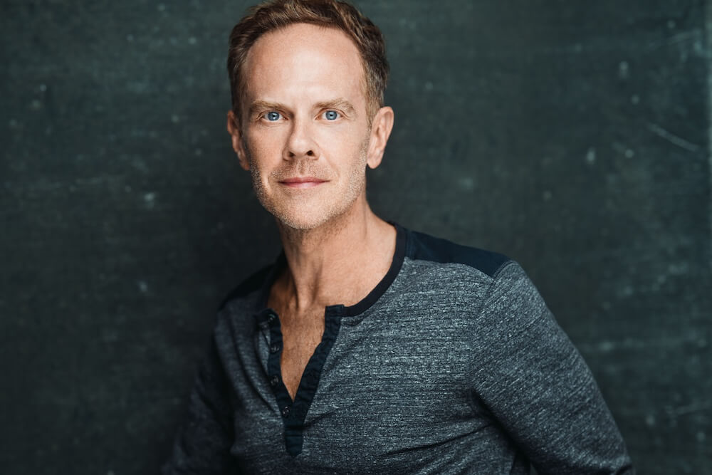 Matthew Neenan, a white man with light short-cut hair and bright blue eyes, stares forward. He wears a grey-ish blue casual button-down shirt.