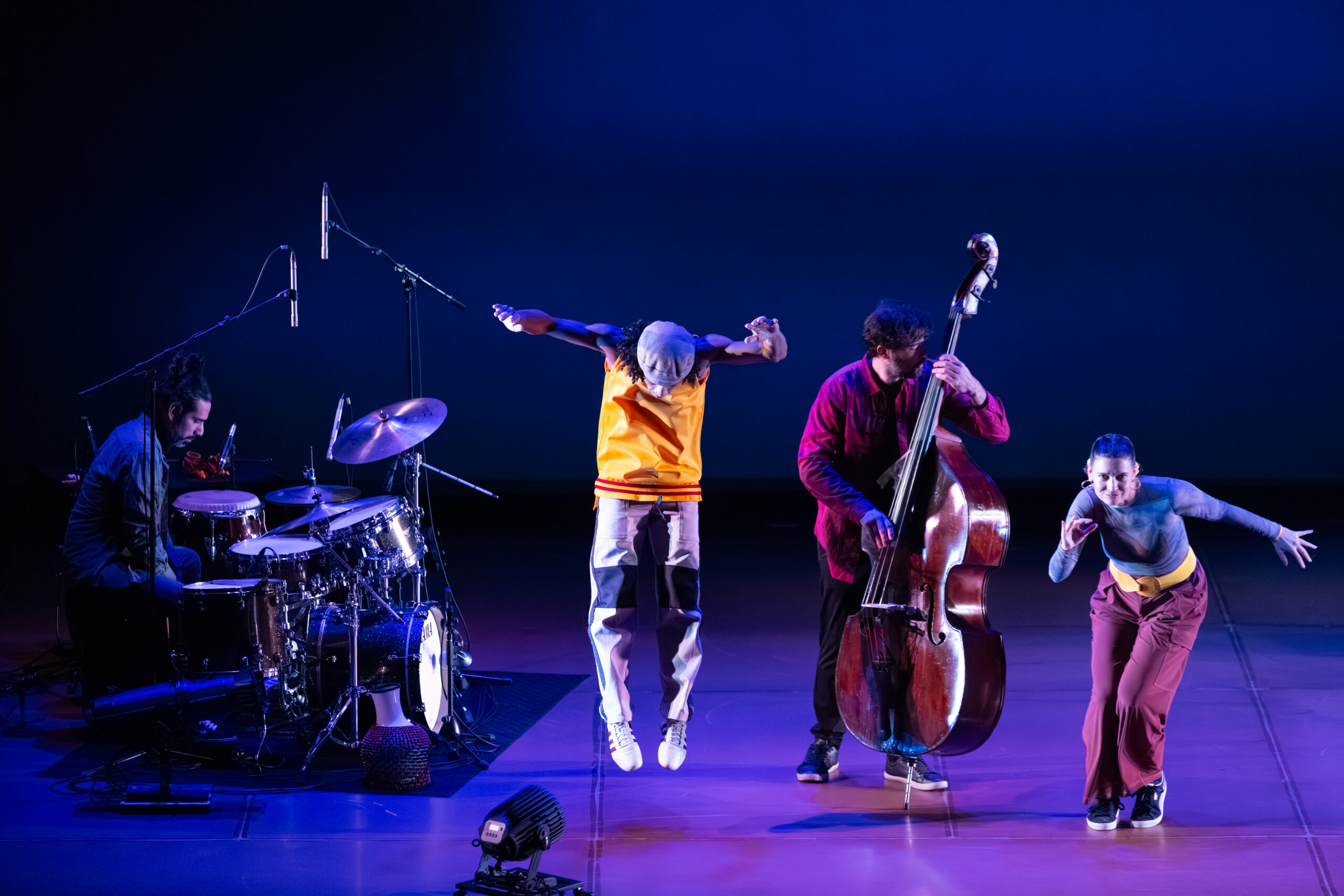 Four performers share a stage lit in deep blue and purple tones. On the left, a musician sits at a drum kit, focused on his instrument, microphones hanging overhead. At center, another performer is captured mid-jump, arms extended overhead and feet dangle above the floor, as if suspended in motion. Beside him, a musician stands playing an upright bass, leaning slightly into the instrument. A small, moveable stage light sits on the floor in front of him. On the far right, a dancer bends forward in a dynamic pose, one arm reaching back and the other curved in front of her, suggesting swift, expressive movement.