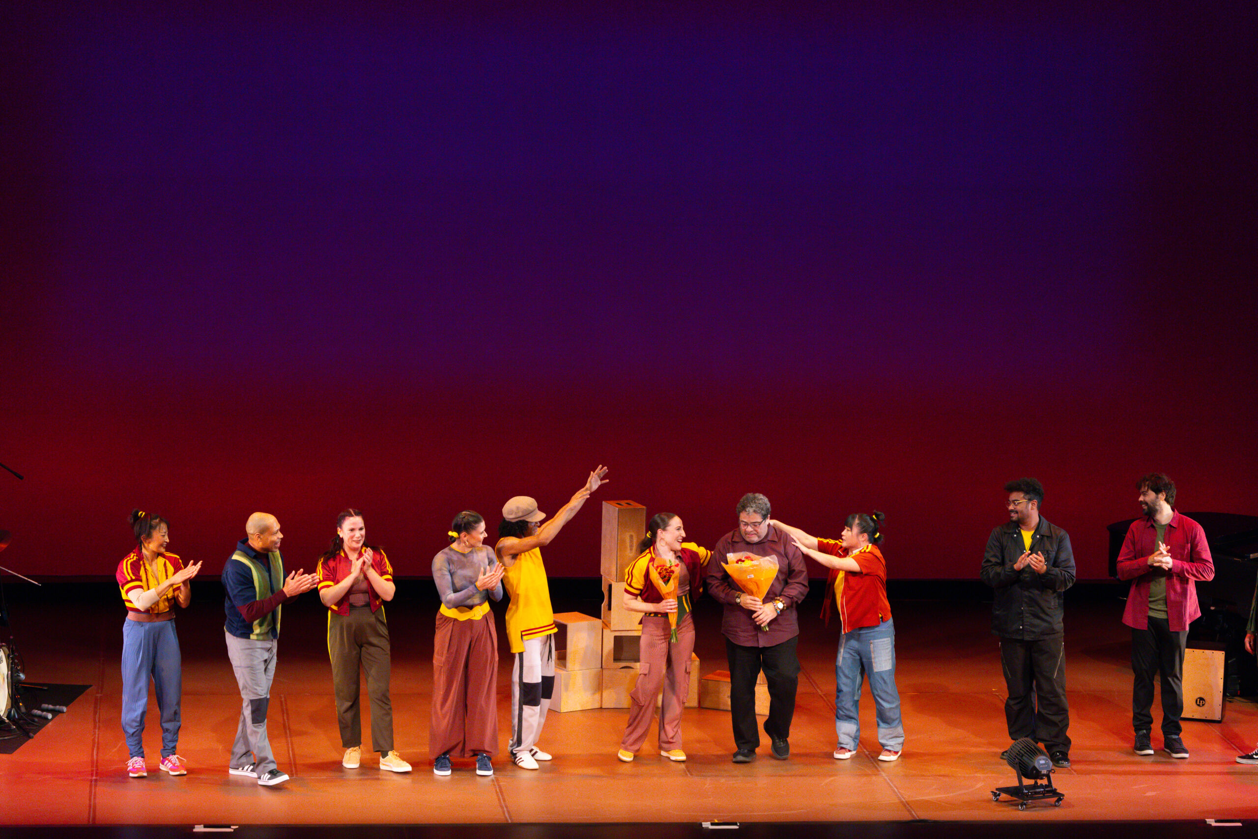 The image shows a group of performers standing in a line onstage, facing the audience. They are positioned in front of a red and blue gradient backdrop. Several people are clapping, as Ephrat Asherie and Arturo O’Farrill each hold a bouquet of flowers center-stage. Wooden boxes are stacked behind the group. Most performers wear casual, colorful clothing, and stage lights and instruments are visible at the edges of the stage.