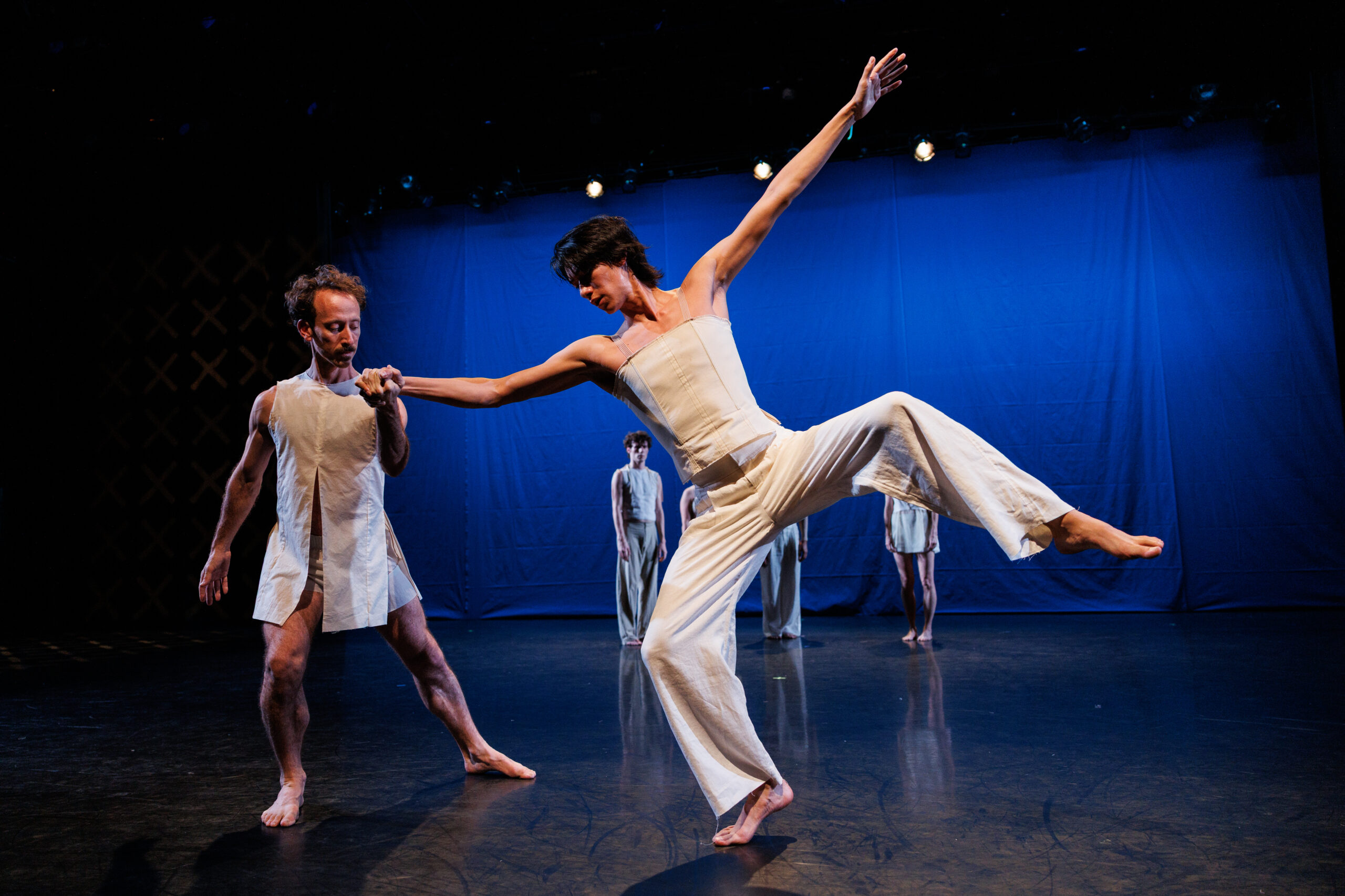 A dancer (Gabriel Bruno Eng Gonzalez), wearing white pants and a sleeveless white top, is balanced on the ball of his right foot. His long arms are extended outwords, and both of his legs are bent. Another dancer (Aaron Loux), also wearing white, lunges on his right leg, and holds Gonzalez´s hand to help him suspend his balancing position. Behind the two of them, in front of a screen the color of deep blue, we can see other dancers standing in the distance, looking at them.