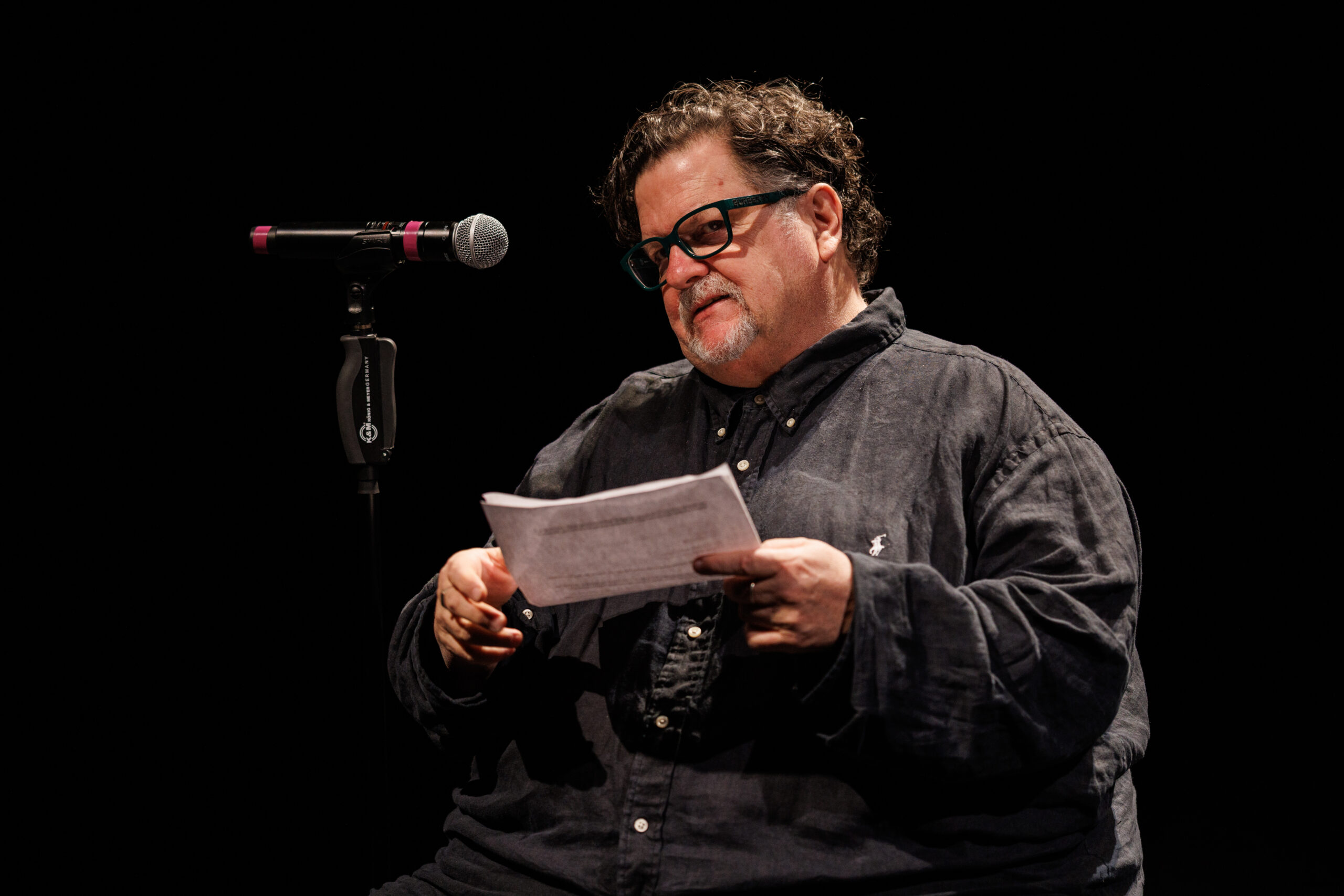 A man (Tere O´Connor) wearing a dark button-down shirt sits in a chair, a microphone in a stand to his right. His head is tilted slightly to one side, and he wears black-rimmed glasses and has a goatee speckled with gray. In his hands he holds some papers, as if he is reading something of importance.