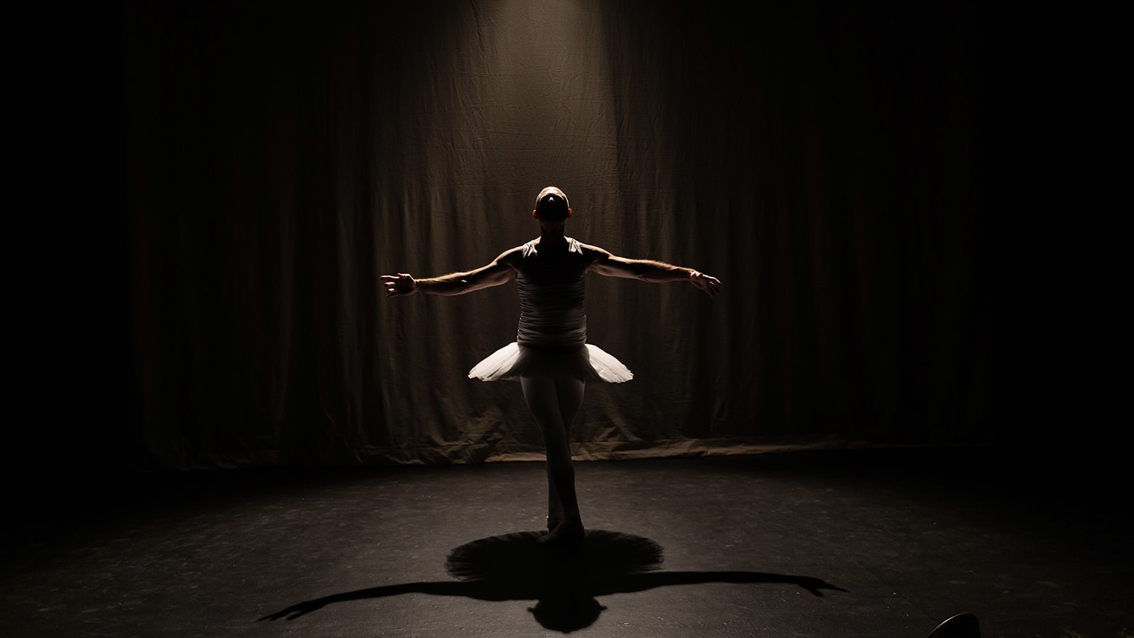 A bald white man wearing white tights and tutu stands center stage in a fifth position soussous with arms stretched sideways, in silhouette on a black marley floor, surrounded by a black backdrop.