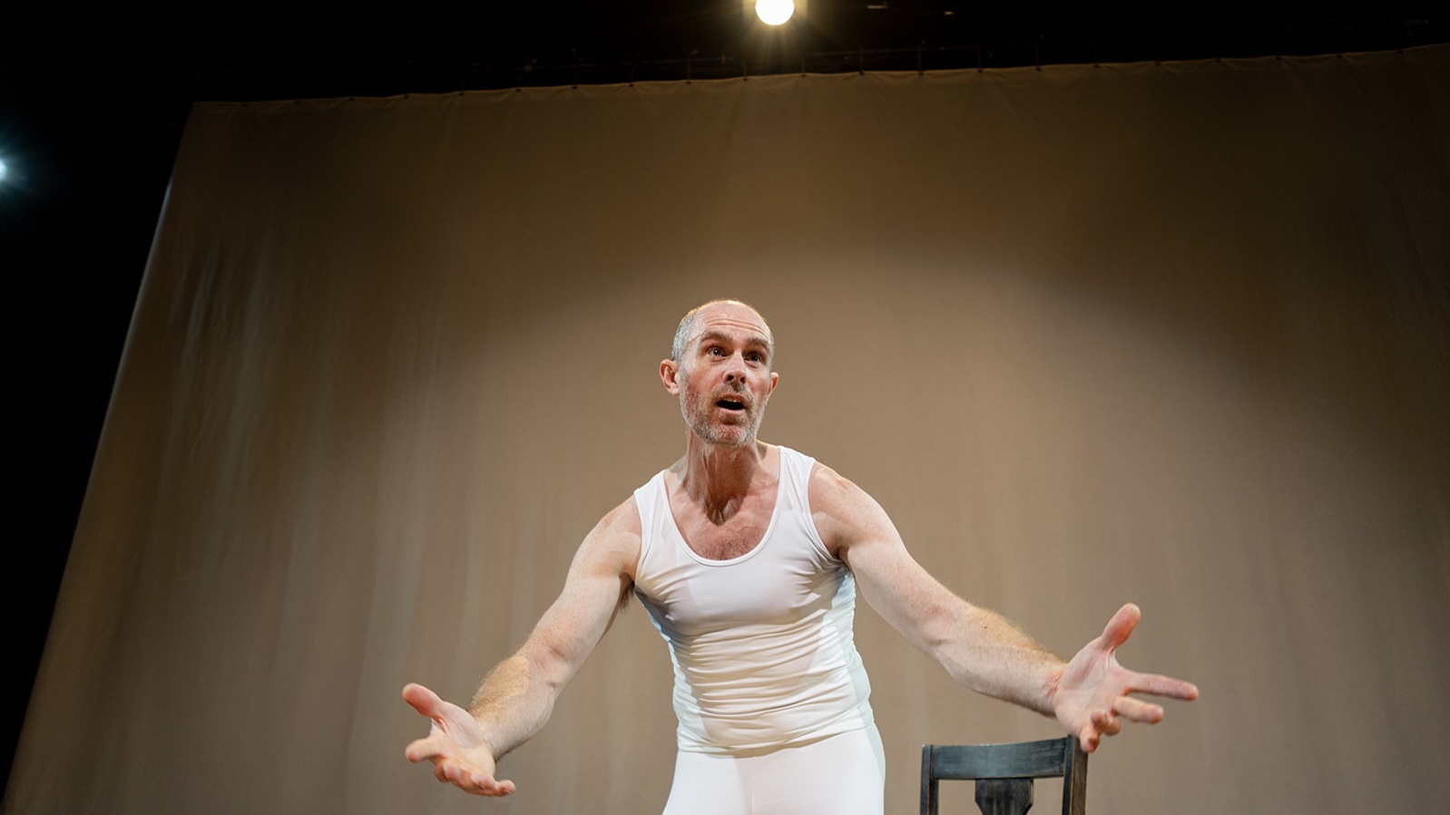 A bald white man wears a tight white tank top and white tights. He leans toward the audience, forehead wrinkled and arms outstretched in a questioning manner.