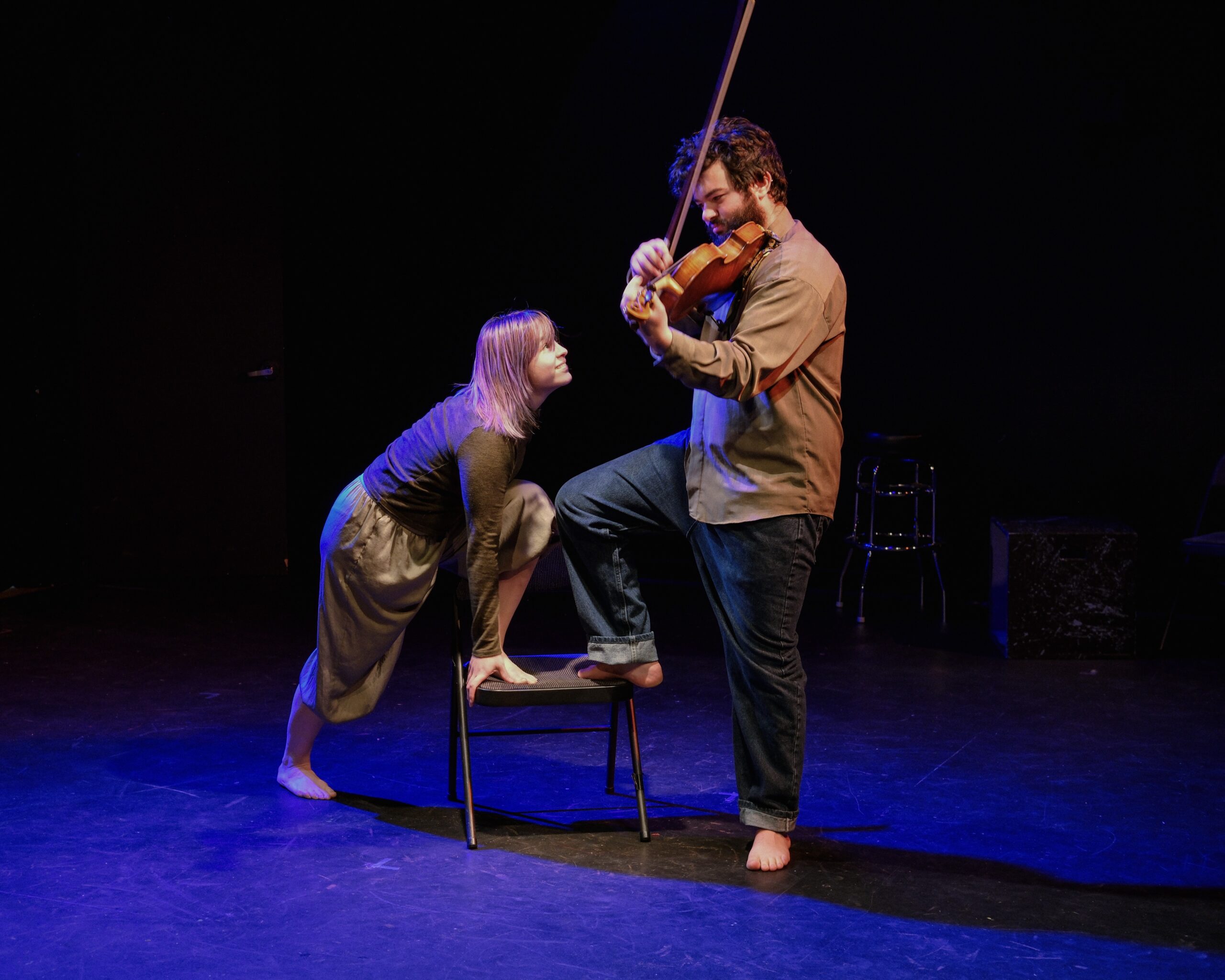 A man wears an untucked brown shirt and jeans. He has one bare foot up on a folding chair and he plays the violin. A woman in shoudler length hair and loosely fitted clothing leans onto the chair with one hand, lunging one bare foot onto it as well. She looks up at the vioilnist smiling impishly.