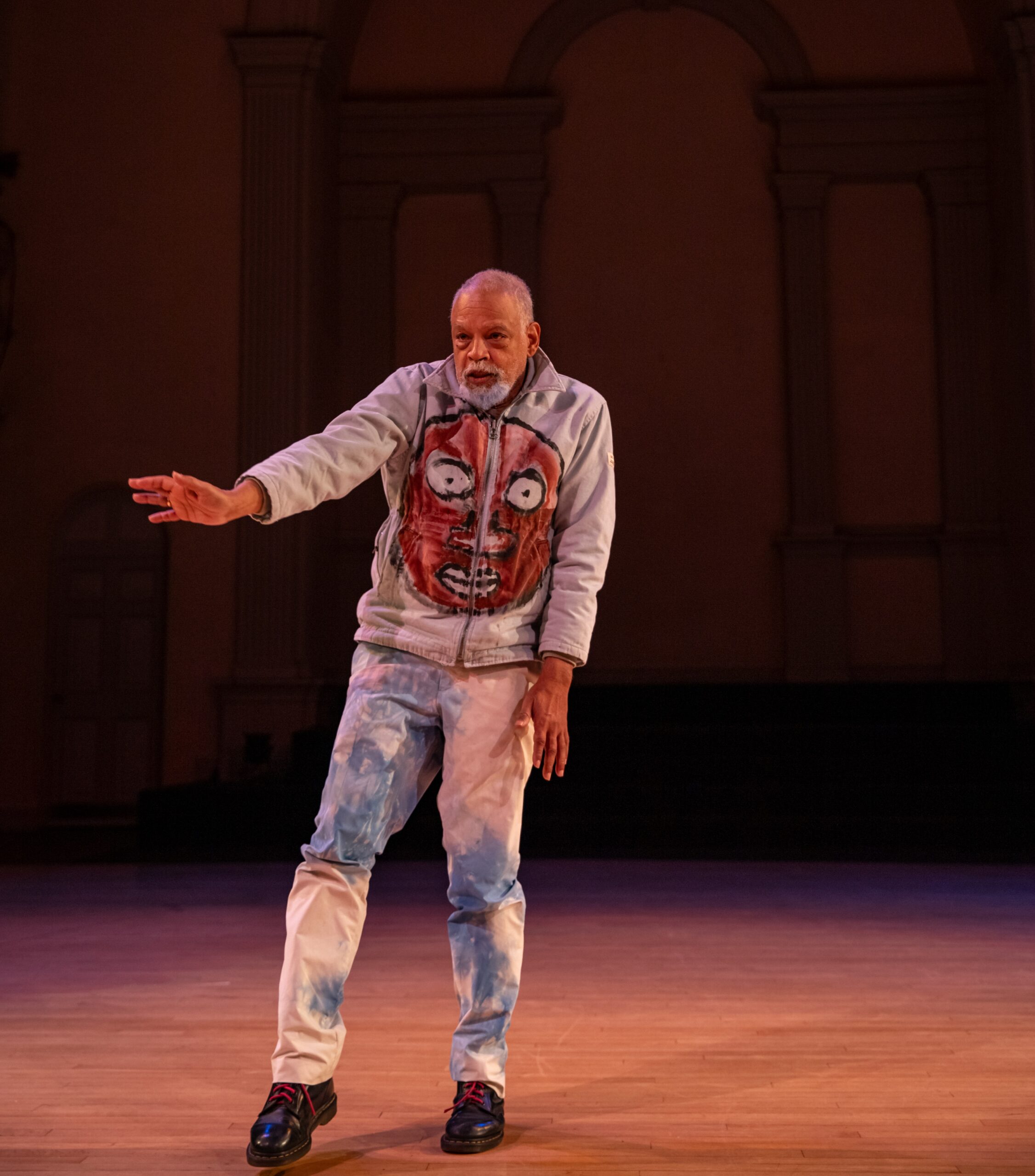 Performer Ishmael Houston-Jones balances on his left foot, his right hand slightly extended forward, an expression of concern on his face. His pants look acid-washed in white and light blue, and on the front of his hoodie is painted a big brown face with wide open eyes.