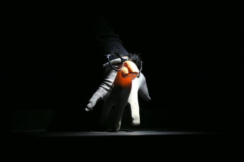 Under a spot light a hand in a white glove is held up as though a small figure is walking. It wears black rimmed glasses with a big plastic nose and fuzzy eyebrows. The arm of the puppeteer is barely visible.